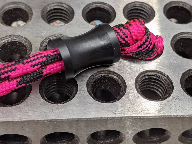 May include: A black cord stopper with a pink and black braided cord threaded through it. The cord stopper is sitting on a metal surface with multiple holes.