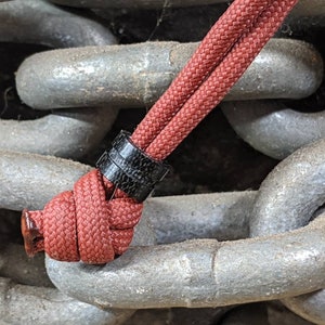 May include: A red and black paracord with a black ring, tied around a chain link. The chain is made of metal and has a silver finish.