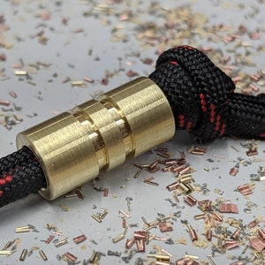 May include: A brass bead with a ridged design, threaded onto a black and red paracord.
