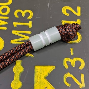 May include: A close-up of a black and brown paracord with a white plastic bead. The paracord is tied in a knot at the end.