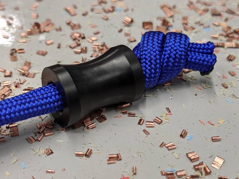 May include: A black plastic cord lock with a blue paracord threaded through it. The cord lock is on a white surface with small copper pieces scattered around it.