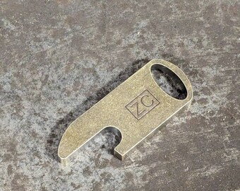 May include: A gold-colored metal bottle opener with a rounded handle and a cut-out for opening bottles. The opener has the letters "ZC" engraved on it.