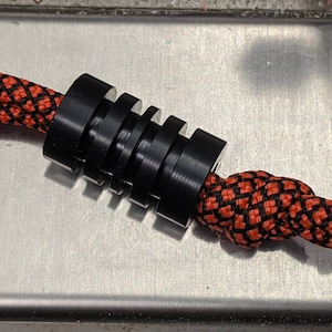 May include: Black cord lock with a ridged design, used for securing cords or ropes. The cord lock is threaded with a black and orange braided cord.