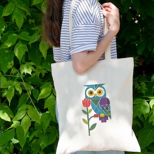 May include: Cream tote bag with an embroidered owl design, featuring yellow eyes, a blue and purple body, and a red flower. The bag is held by a person wearing a striped top, with green leaves in the background.