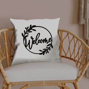 Pode incluir: Uma almofada quadrada branca com a palavra "Welcome" em preto, em fonte cursiva, circundada por uma grinalda floral preta. A almofada está numa cadeira de vime com uma almofada branca, contra um fundo neutro.