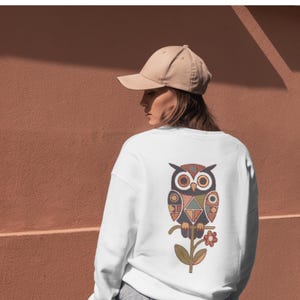 Peut inclure: Sweat-shirt blanc orné d'un motif de hibou coloré dans le dos. Le hibou est représenté avec des formes géométriques dans des tons de marron, orange, bleu et vert. Une casquette de baseball beige est portée.