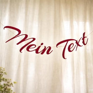 Può includere: Uno striscione con le parole "Mein Text" in un carattere corsivo bordeaux, appeso su uno sfondo dai toni neutri. Lo striscione è sospeso da un filo sottile, creando un'esposizione semplice ed elegante.