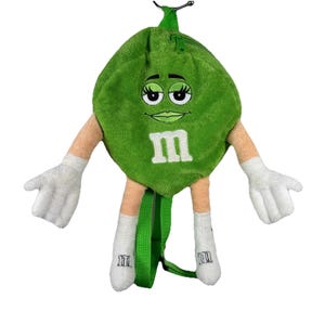 May include: A green plush backpack shaped like a candy character, featuring a white "M" on the front. The backpack has arms with white gloves, and legs with white socks. The straps are green, and the zipper is black.