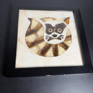 May include: A square wall art piece featuring a stylized raccoon face crafted from layered brown and cream-colored butterfly wings. The artwork is set against a textured off-white background and enclosed within a simple black wooden frame.