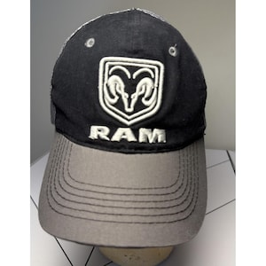 May include: Black and gray baseball cap with the word "RAM" in white. The cap features a white emblem of a ram's head on the front. The brim is gray, and the back is mesh.