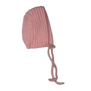 May include: A light pink knitted baby bonnet with vertical ribbing. The bonnet has long, knitted ties for securing under the chin. The soft, pastel color and simple design suggest a focus on comfort and warmth for infants.