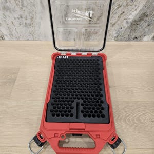 May include: Open red and black Milwaukee Packout storage case. The clear lid reveals compartments, while the interior features a black foam insert. The case is designed for organization and storage, with a focus on durability and accessibility.