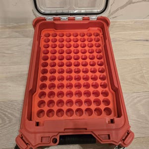 May include: A red plastic storage case with a clear lid, open to reveal a grid of circular holes. The case is designed to hold small items, with a handle and wheels for easy transport. The case is empty.