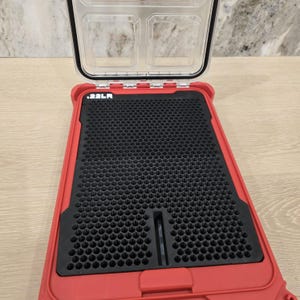 May include: Open red storage case with a clear lid. The interior features a black, textured insert with a honeycomb pattern. The text ".22LR" is visible on the case. The case is designed to hold small items.