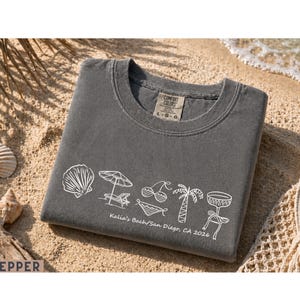 May include: A grey Comfort Colors t-shirt featuring white beach-themed illustrations, such as a seashell, parasol, sun lounger, cherries, bikini, palm tree, and a drink. The text "Kalia's Bach/San Diego, CA 2026" is printed below the illustrations.