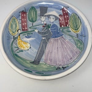 Könnte beinhalten: Ein Keramikteller mit handbemaltem Design eines Paares, das in einem Park spaziert. Der Teller hat einen blauen Hintergrund mit grünen Bäumen, roten Gebäuden und einem gelben Tisch mit einer weißen Tischdecke. Das Paar ist in Kleidung aus dem 19. Jahrhundert gekleidet und hält sich an den Händen.