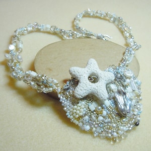 May include: A handcrafted necklace featuring a heart-shaped pendant adorned with white, silver, and gold beads, and a white starfish. The necklace has a beaded chain with a silver clasp. The jewelry is set on a light beige surface.