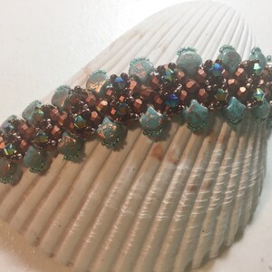 May include: A beaded bracelet featuring a turquoise and copper colour scheme. The bracelet is crafted with small, faceted beads and larger, irregular beads. The bracelet is displayed on a white seashell.