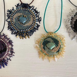 May include: Four handmade beaded necklaces with circular pendants. The pendants feature intricate beadwork in various colors, including purple, blue, gold, and silver, with a central shell-shaped bead. Each necklace has a different cord color.