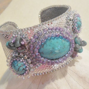 Op de afbeelding: Een zilveren en turquoise kralen armband met een geweven gaasontwerp. De armband heeft drie turquoise stenen en paarse kralen.