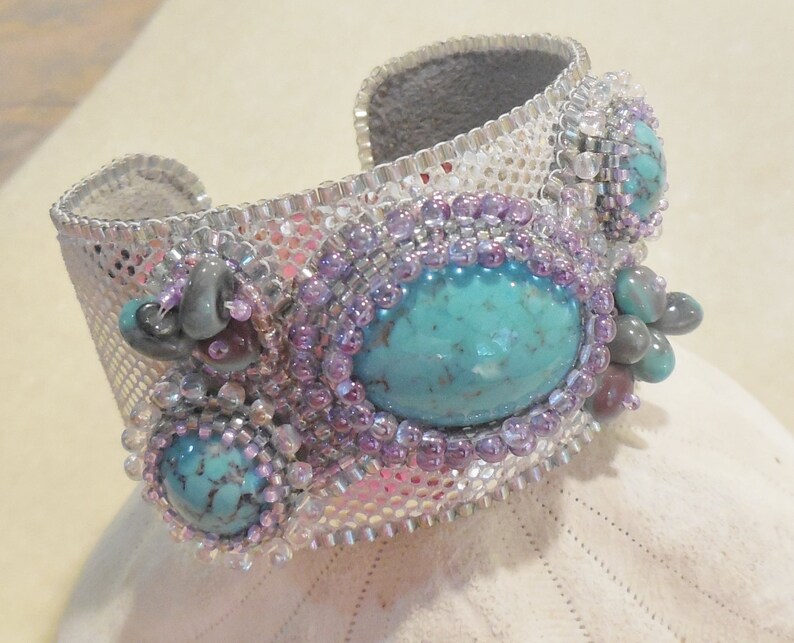 Op de afbeelding: Een zilveren en turquoise kralen armband met een geweven gaasontwerp. De armband heeft drie turquoise stenen en paarse kralen.
