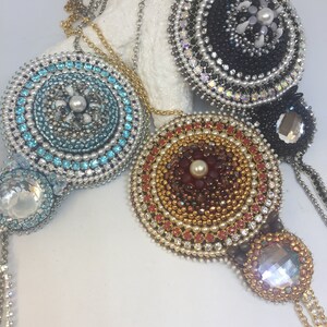May include: Four beaded pendant necklaces in various colors. The pendants are round with intricate beadwork and crystal accents. Colors include light blue, black, gold, and red. Each necklace has a chain and a crystal pendant at the bottom.