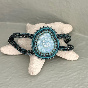 May include: A beaded bracelet featuring a central iridescent light blue ammonite-shaped cabochon. The focal piece is surrounded by concentric rings of small turquoise, dark blue, and metallic beads, with a band made of dark, shimmering seed beads.