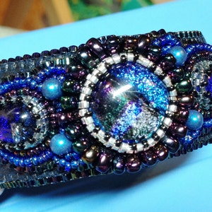 Op de afbeelding: Een zwarte leren armband met een grote blauwe en zilveren glazen cabochon, omringd door donkerpaarse, zilveren en blauwe kralen.