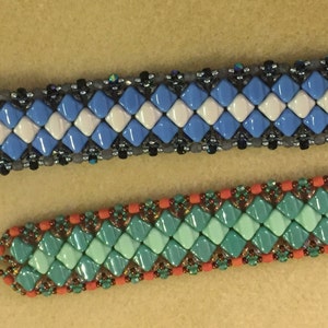 May include: Two beaded bracelets. The top bracelet features a blue and white diamond pattern with a dark gray border. The bottom bracelet has a turquoise and green diamond pattern with an orange border. Both bracelets have a clasp.