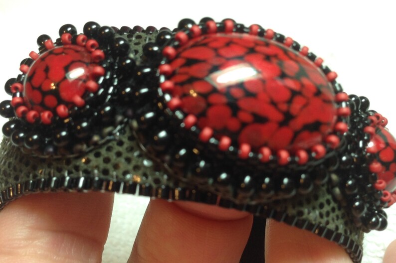 Op de afbeelding: Een zwarte en rode kralen armband met drie rode en zwarte gepatroneerde cabochons. De armband is gemaakt met een zwarte mesh basis en heeft een zwarte kralen rand.