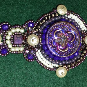 May include: A purple and white beaded brooch with a large iridescent purple button in the center. The button has a flower design.
