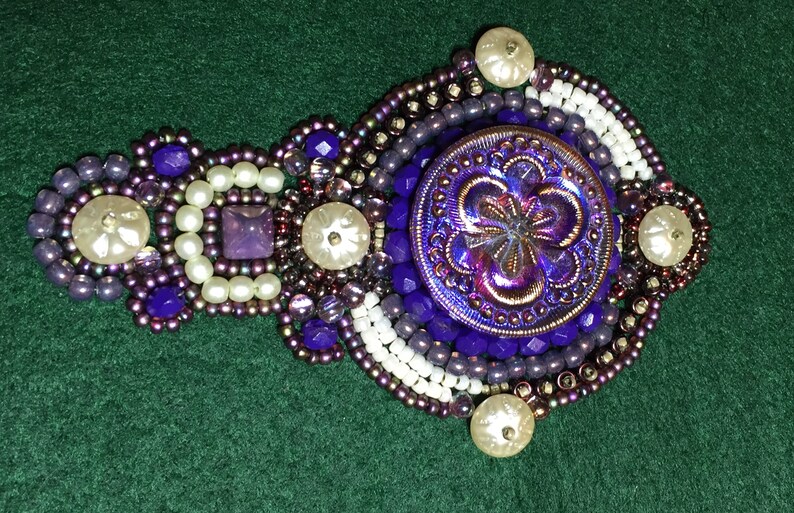 May include: A purple and white beaded brooch with a large iridescent purple button in the center. The button has a flower design.