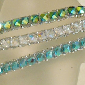 Può includere: Tre braccialetti con perline argentate e dettagli in cristallo. Il braccialetto superiore ha perline verdi iridescenti, il braccialetto centrale ha perline di cristallo trasparenti e il braccialetto inferiore ha perline blu iridescenti.