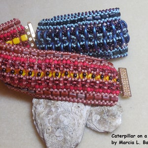 Könnte beinhalten: Zwei Perlenarmbänder, eines rot, gelb und braun, das andere blau und lila, mit goldenen Verschlüssen. Die Armbänder liegen auf einem hellgrauen Stein. Der Text "Caterpillar on a Tile Road by Marcia L. Balonis" befindet sich unterhalb der Armbänder.
