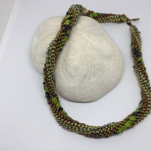 May include: A green, brown, and silver beaded necklace with a twisted design. The necklace is displayed on a white surface.