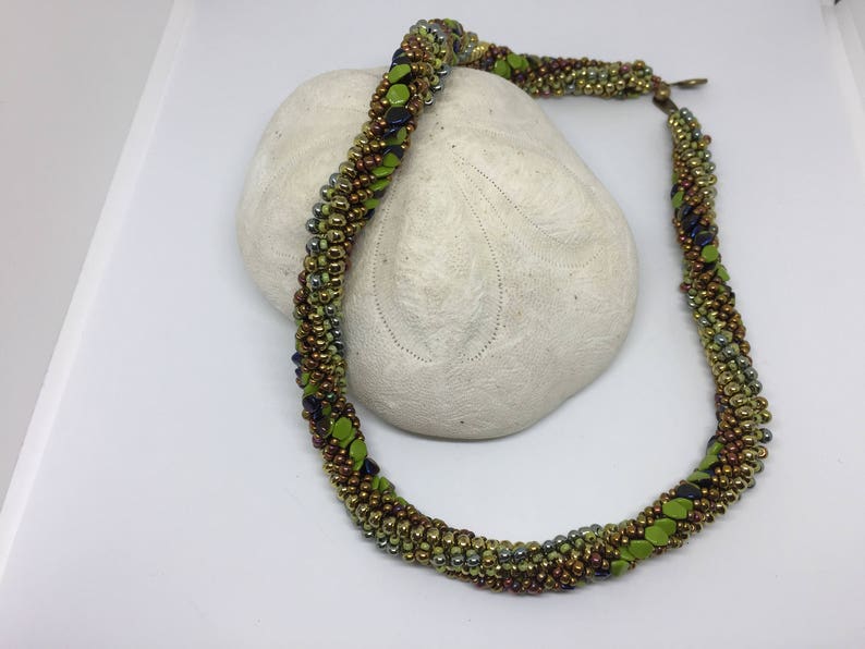 May include: A green, brown, and silver beaded necklace with a twisted design. The necklace is displayed on a white surface.