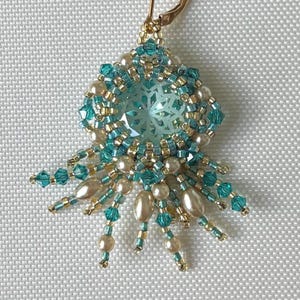 May include: A handmade earring featuring a central, faceted, light blue gemstone surrounded by teal and gold beads. The design includes teardrop-shaped pearls and teal crystal accents, creating a delicate, floral-like pattern. The earring has a gold-colored clasp.