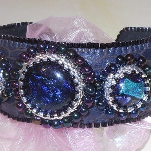 Op de afbeelding: Een zwarte leren armband met drie iriserende blauwe glazen cabochons, gezet in zilveren kralencirkel. De armband is versierd met zwarte en zilveren kralen.