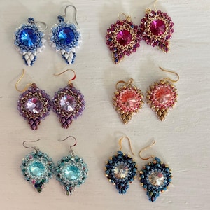 May include: A collection of handmade beaded earrings in various colors. The earrings feature large, faceted crystal center stones surrounded by intricate beadwork in shades of blue, purple, pink, and teal. Each pair has a unique design and gold or silver ear wires.