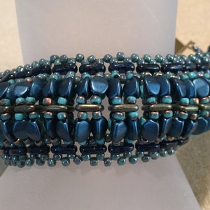 May include: A blue and turquoise beaded bracelet with a metallic finish. The bracelet has a geometric pattern and is made with various sizes of beads.