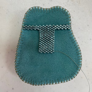 Puede incluir: Primer plano de una bolsa de tela azul turquesa con un diseño de cuentas plateadas cosido en la parte delantera. La bolsa está rodeada por un ribete dorado.