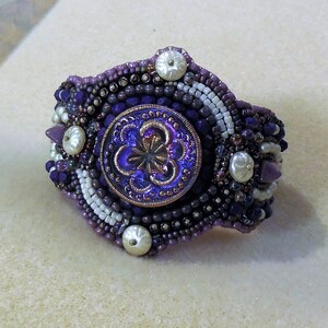 May include: A purple and white beaded bracelet with a large iridescent purple button in the center. The button has a floral design.