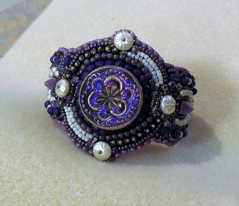 May include: A purple and white beaded bracelet with a large iridescent purple button in the center. The button has a floral design.