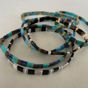 May include: A set of four colorful beaded bracelets in shades of blue, turquoise, black, white, and gold. The bracelets are stacked on top of each other, creating a layered effect.