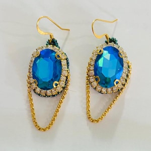 May include: A pair of gold earrings with a large blue oval-shaped gemstone surrounded by smaller clear rhinestones. The earrings have a dangling chain.