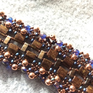 May include: A close-up of a beaded bracelet with copper and blue beads. The bracelet is made with a woven pattern of copper and silver squares.