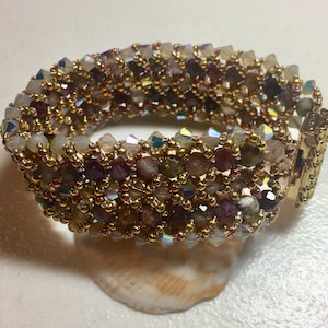 May include: A beaded bracelet featuring a mix of colorful gemstones and gold-colored beads. The bracelet has a woven design with a gold clasp. The gemstones include shades of green, purple, and clear crystals. The bracelet is resting on a white surface.