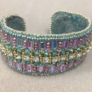 May include: A beaded cuff bracelet with a turquoise base and a band of pink, blue, and clear beads. The band is accented with a row of clear rhinestones.
