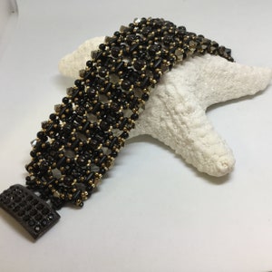 Op de afbeelding: Een zwarte en gouden kralen armband met een geometrisch ontwerp. De armband is gemaakt met kleine, ronde kralen en heeft een sluiting.