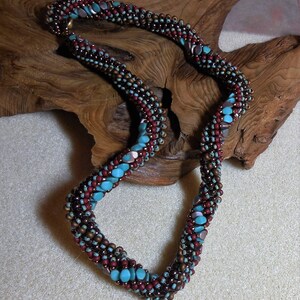 May include: A brown, red, and turquoise beaded necklace with a twisted design. The necklace is made of small, round beads and is displayed on a wooden surface.